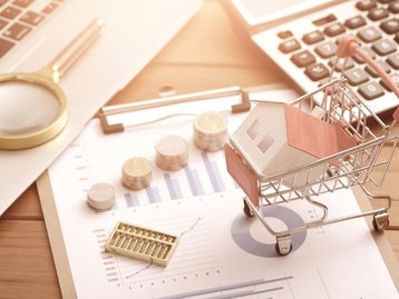 Kosten einer Immobilien-Kapitalanlage kalkulieren Einkaufswagen mit Hausmodell, Münzen, Diagrammen und Taschenrechner zur Kalkulation einer Immobilieninvestition